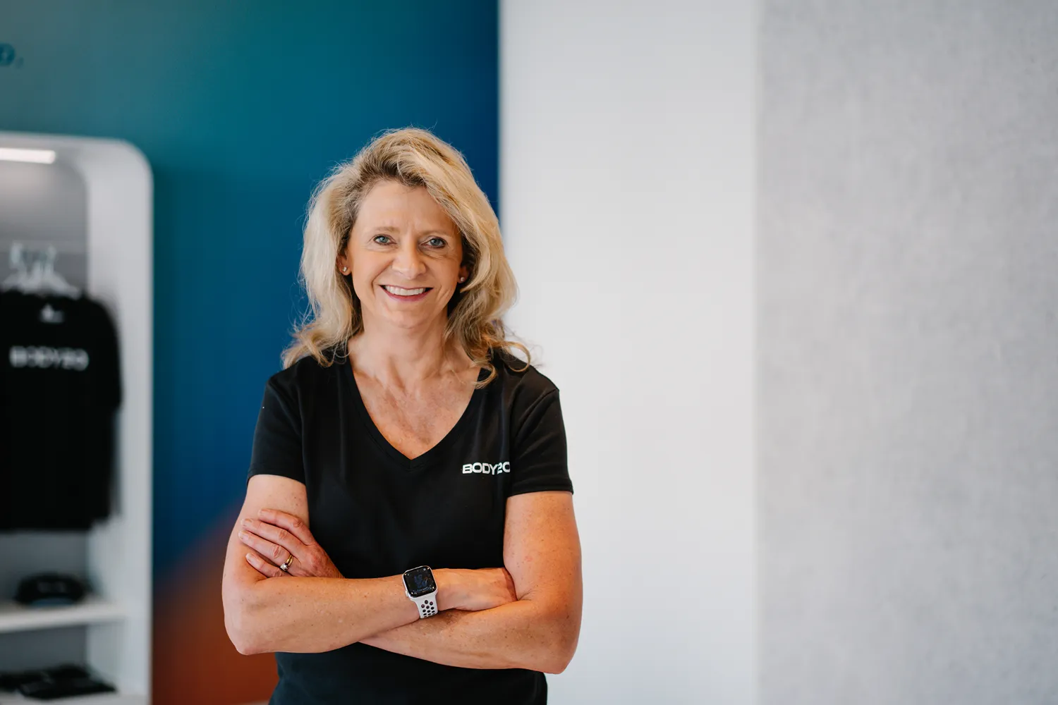 Confident business owner smiling with arms crossed, standing inside a modern fitness studio wearing branded apparel—professional branding headshot with a clean, minimal background.