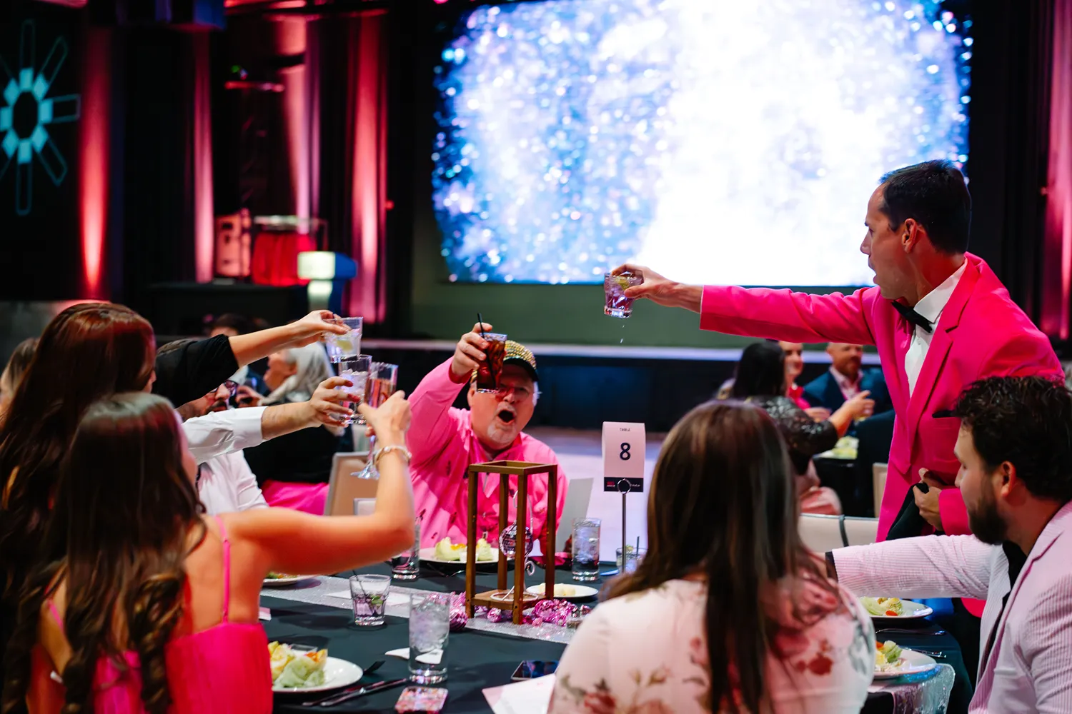 Guests wearing pink and white outfits raising glasses in a celebratory toast around a decorated table at a StatLab award ceremony in Dallas.