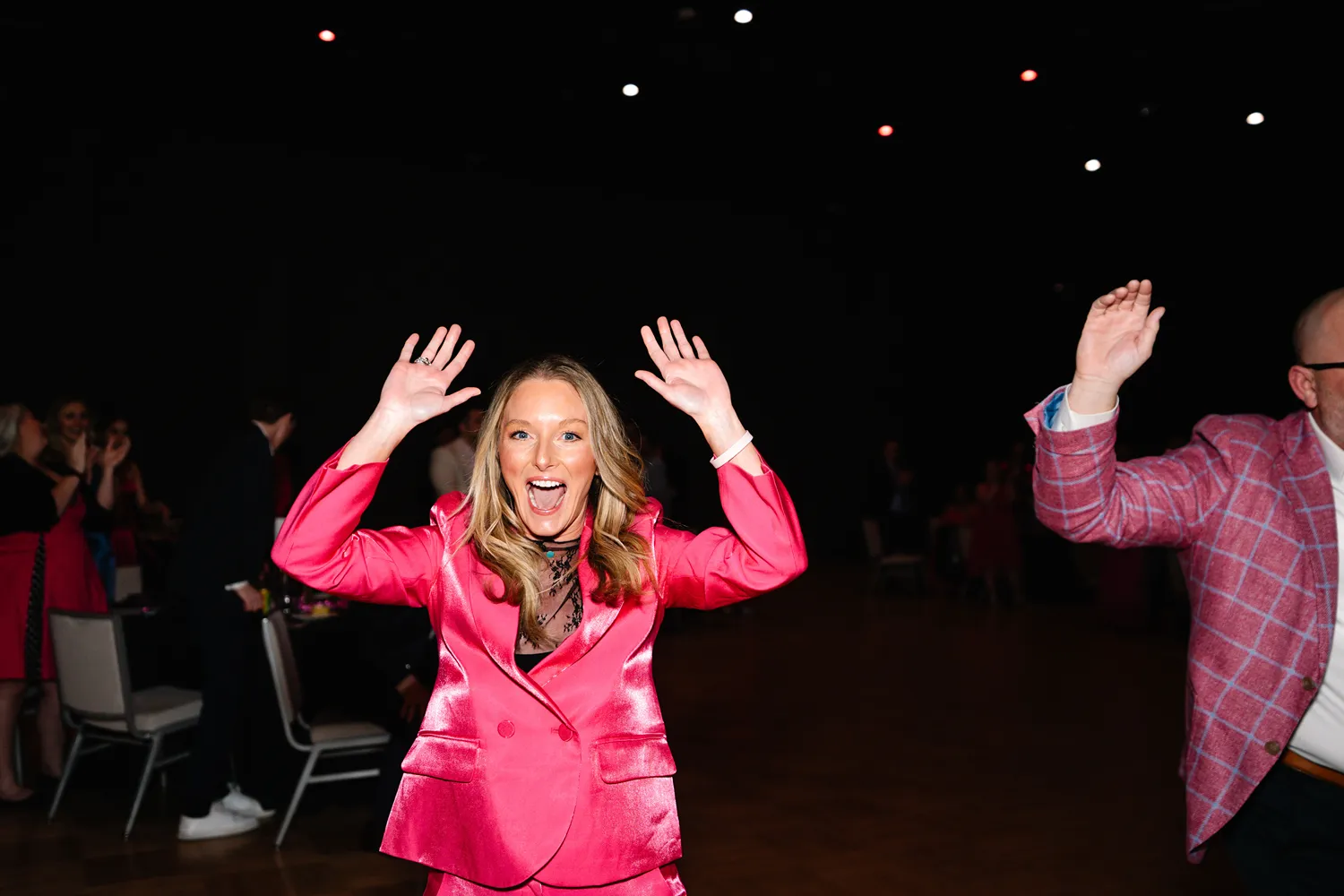 A woman in a bright pink suit raises her hands and smiles enthusiastically on the dance floor at a StatLab event.