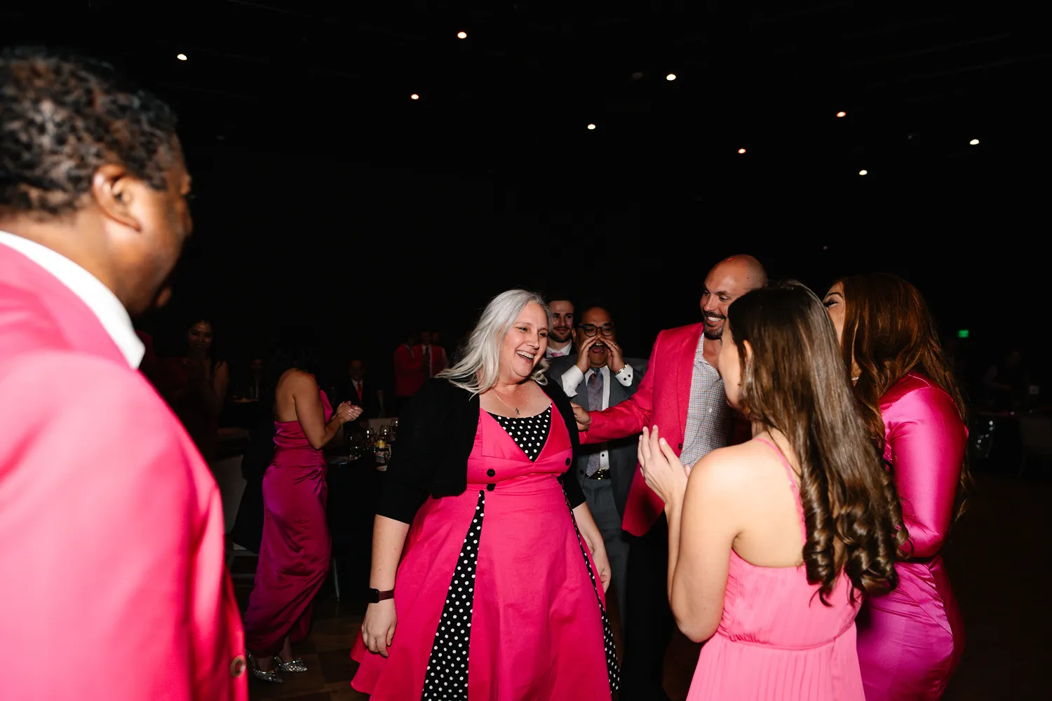 A group of people in pink attire gather and applaud a smiling woman in a polka-dot dress during candid moments at a Dallas corporate awards celebration.