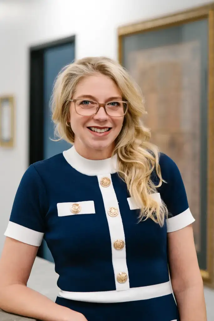 Smiling woman in a navy and white dress with gold buttons, posing confidently in a modern office setting with framed artwork in the background.