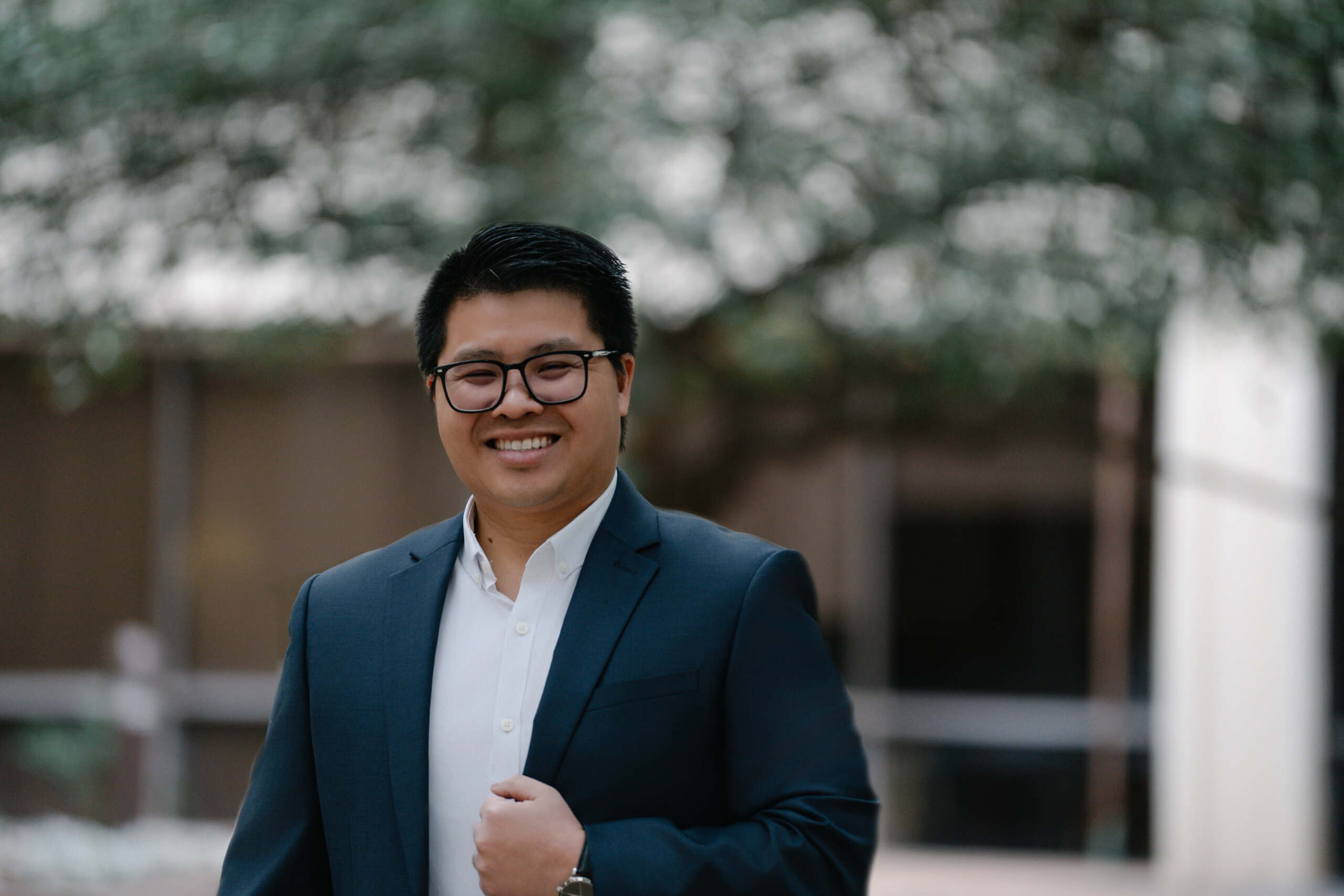 Professional traditional headshot of a man in a navy suit and white shirt, smiling outdoors with a softly blurred background—ideal for corporate profiles, LinkedIn, and company websites.