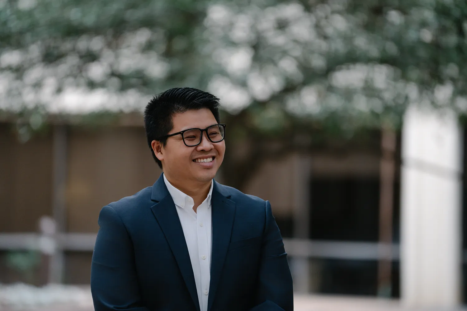 Branding photo of a man in a navy suit and glasses, smiling and looking off-camera outdoors—this relaxed, approachable image style is ideal for business websites, social media, and marketing materials, going beyond a traditional headshot to showcase personality and brand story.
