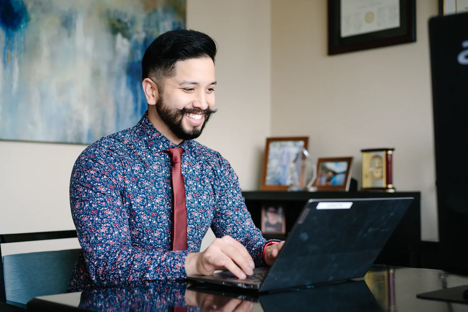 A smiling man in a floral shirt and red tie works on a laptop at his desk in a professional office setting. Framed photos, awards, and a diploma are visible in the background, creating a warm and welcoming atmosphere.
