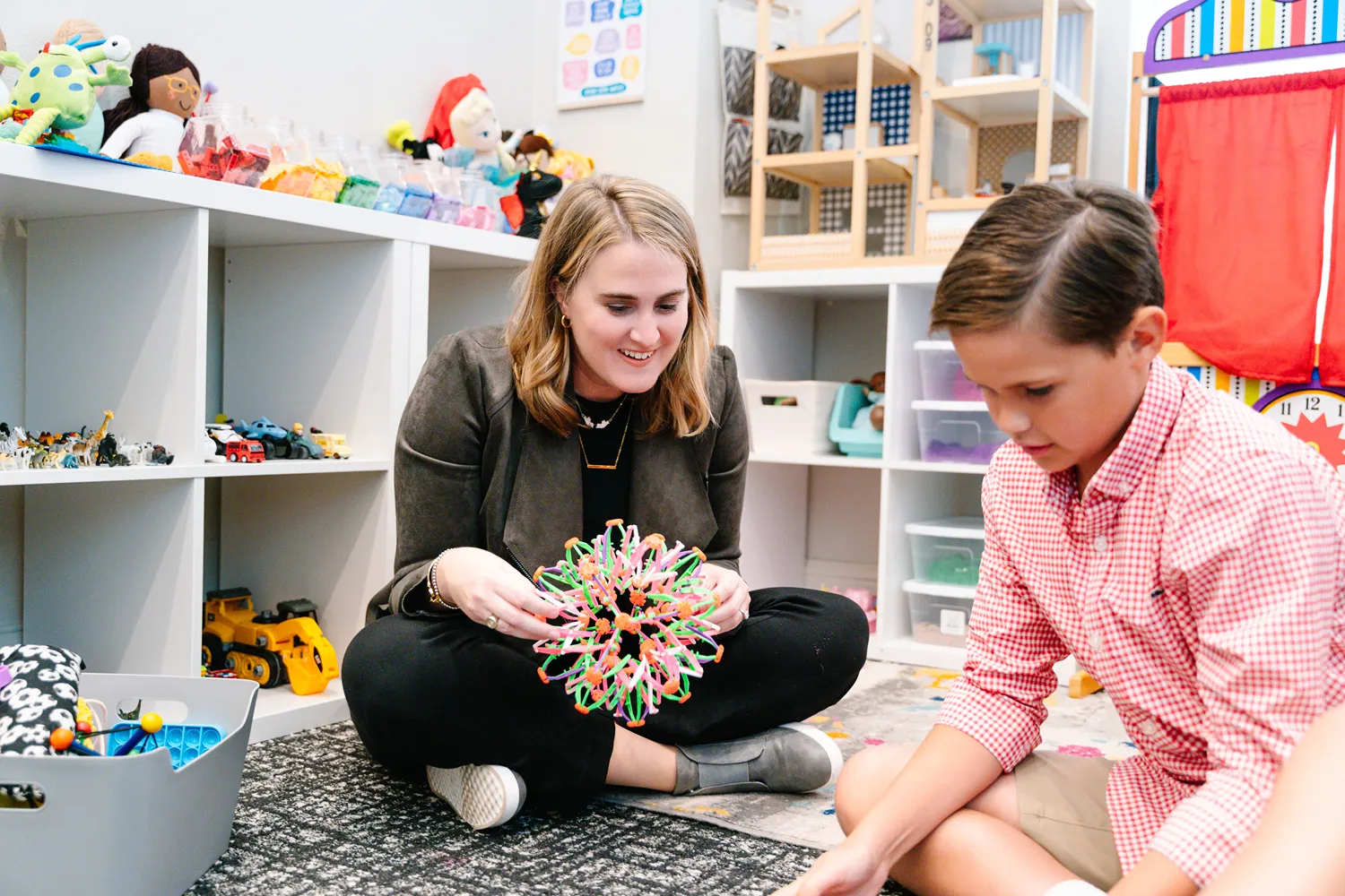 Female therapist smiling while playing with a young client in a colorful, toy-filled therapy room, showcasing a warm and engaging brand atmosphere.
